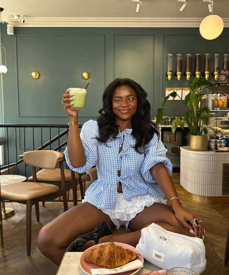 Uma mulher sorridente sentada em um café chique, segurando um copo de bebida verde. Ela usa uma blusa xadrez azul e branca, shorts brancos e mocassins pretos. Um croissant e uma bolsa de maquiagem estão na mesa.