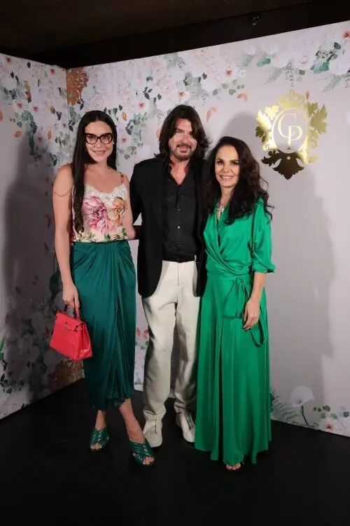 Anne Williams vestindo blusa floral com renda e saia verde bandeira, Cris Porto de verde e Stephane Rolland de terno preto e calça off-white em fundo com papel de parede com logo da Cris Porto joalheria e flores