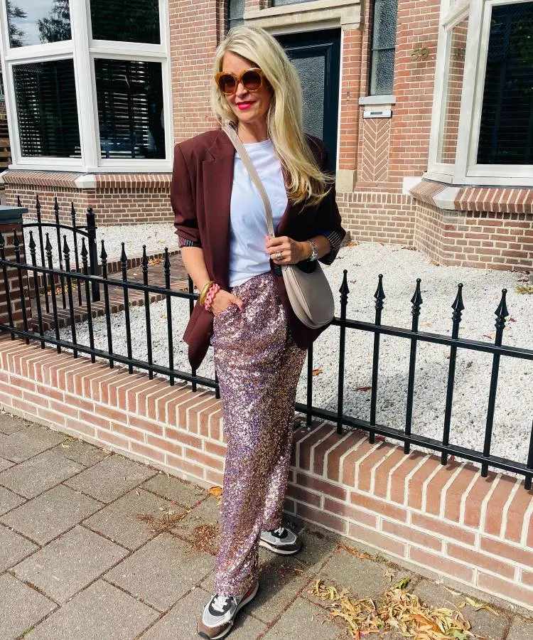 A mulher posa confiante em frente a uma casa de tijolos, vestindo calça brilhante de paetês rosa, camiseta branca e blazer vinho. Óculos de sol grandes, bolsa bege e tênis esportivos completam o look elegante e casual.