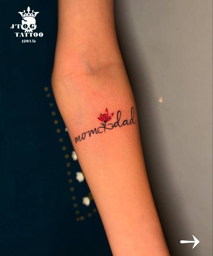 A imagem mostra uma tatuagem no antebraço de uma pessoa com as palavras "mom & dad" em uma fonte cursiva preta, onde o "e" de "mom" é substituído por uma flor de lótus vermelha. O fundo é cinza escuro. No canto superior esquerdo há um logotipo e um texto em branco.







