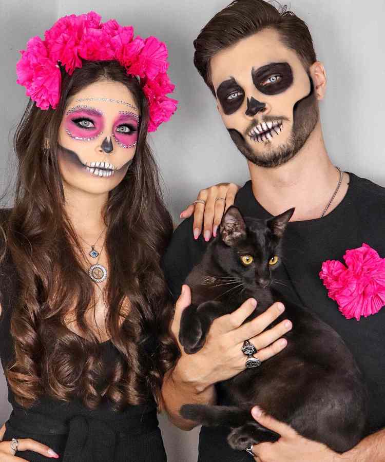 Fantasia de casal para o Halloween: Caveira Mexicana. Ambos usam roupa preta. A mulher usa maquiagem rosa de caveira e flores no cabelo. O homem usa maquiagem preta 