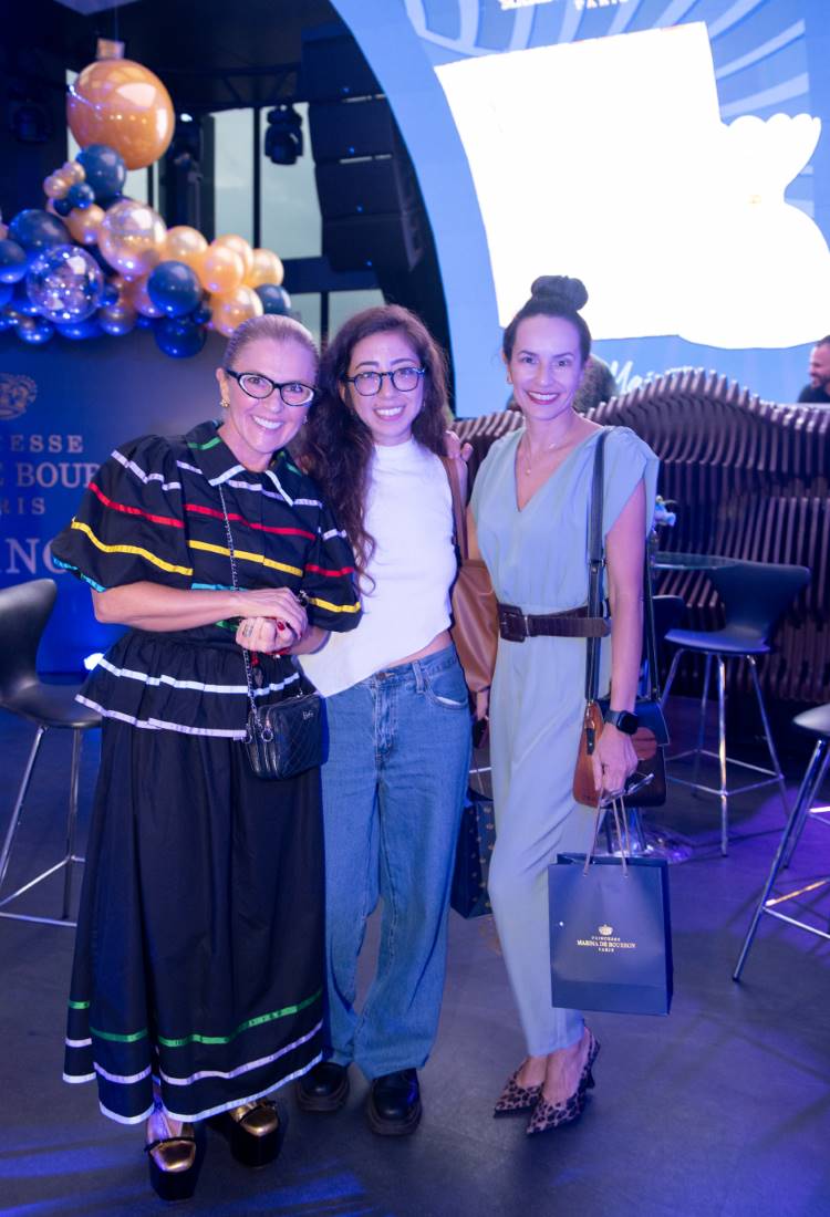 Três mulheres posam sorridentes em frente a uma decoração temática azul, com balões dourados e azuis. O evento celebra "Princesse Marina de Bourbon Paris". Cada uma tem um estilo distinto, com roupas elegantes. Ambiente sofisticado e festivo.