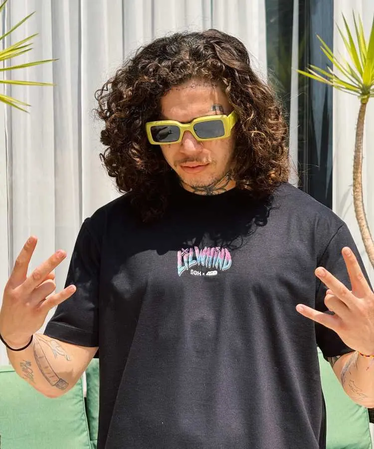 A imagem mostra Whindersson Nunes com cabelos longos e cacheados, usando óculos de sol amarelos e uma camiseta preta com estampa. Ele faz gestos com as mãos e está ao ar livre, com plantas e cortinas ao fundo.