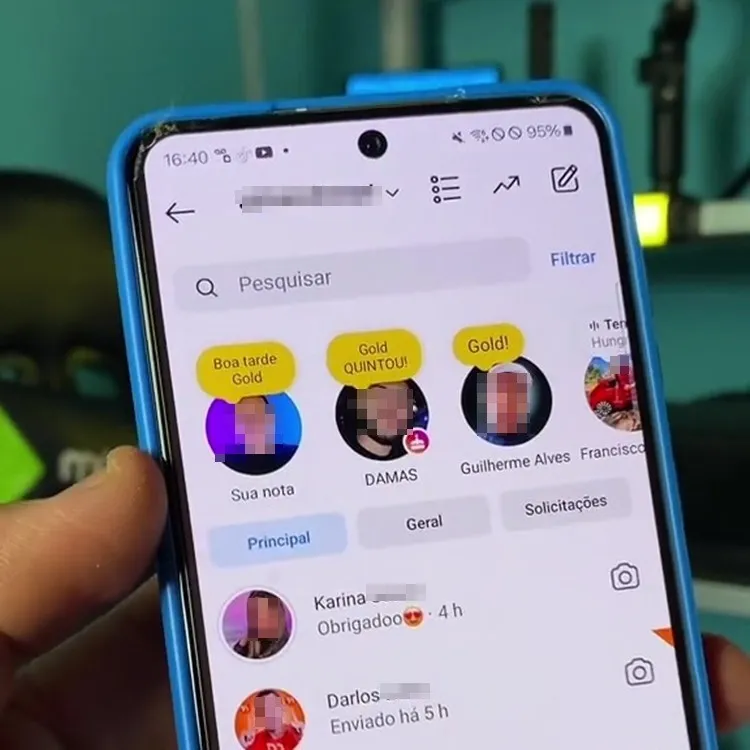 A imagem mostra a tela de um smartphone exibindo a interface de mensagens diretas do Instagram. No topo, há ícones de stories de diferentes usuários, alguns com etiquetas douradas que dizem "Boa tarde Gold", "Gold QUINTOU!" e "Gold!". Abaixo, há uma barra de pesquisa e filtros, seguida por três abas: "Principal", "Geral" e "Solicitações". Duas mensagens recentes são visíveis, uma de "Karina" com a resposta "Obrigadoo?" enviada há 4 horas e outra de "Darlos" com "Enviado há 5 h". A mão de uma pessoa segura o telefone, que está em uma capa azul. Ao fundo, é possível ver um ambiente desfocado com alguns objetos.


