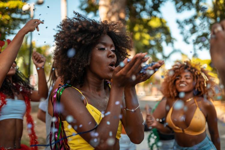 Mulher soprando confetes em um ambiente festivo ao ar livre, cercada por amigos comemorando. Todos vestem roupas coloridas e brilhos, representando alegria e energia do Carnaval. Árvores e luz natural complementam o cenário vibrante e descontraído da celebração.