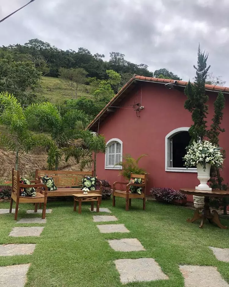 Fachada de casa vermelha com janelas brancas em área rural. Plantas em vasos, palmeiras e pinheiro decoram o jardim gramado. Há caminhos de pedras retangulares e um jogo de sofá, poltronas e mesinha, com almofadas estampadas de costela de adão