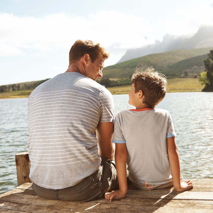 Um homem e um menino sentam-se lado a lado em um píer de madeira, admirando um lago sereno com montanhas ao fundo. Ambos sorriem enquanto conversam, simbolizando companheirismo, confiança e momentos especiais compartilhados ao ar livre.