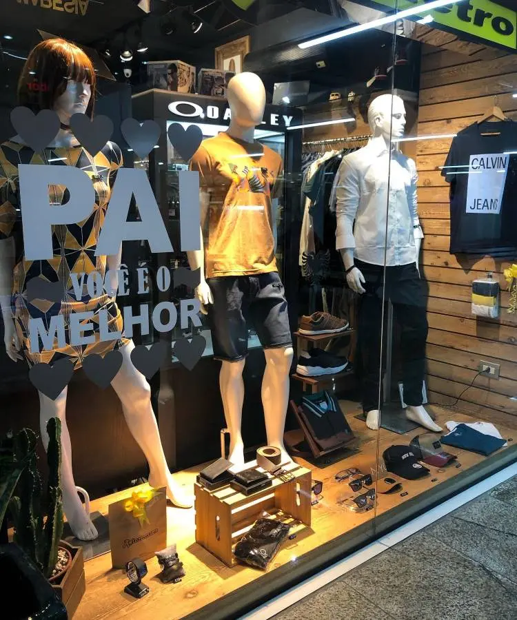 A imagem exibe uma vitrine de loja com três manequins apresentando roupas masculinas. Há um adesivo na vitrine com a frase "PAI VOCÊ É O MELHOR" e corações decorativos, sugerindo uma promoção para o Dia dos Pais.