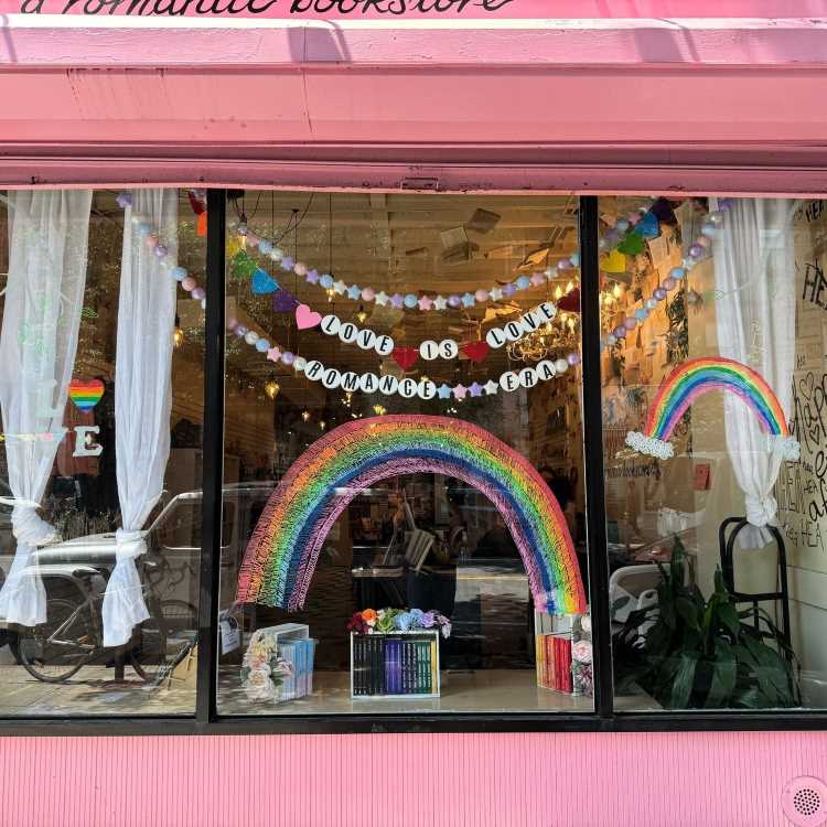 Fachada de livraria rosa com vitrine decorada. Arco-íris central brilhante e bandeirolas com letras "Love is Love" destacam diversidade e orgulho. Livros coloridos e flores enfeitam a base. Cortinas brancas, plantas e bicicleta criam ambiente acolhedor e vibrante.