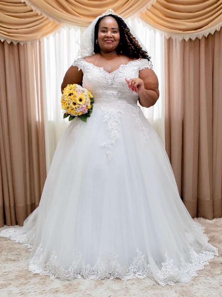 Mulher de pele negra usando vestido de noiva bordado com rendas e segurando buquê de flores amarelas. 