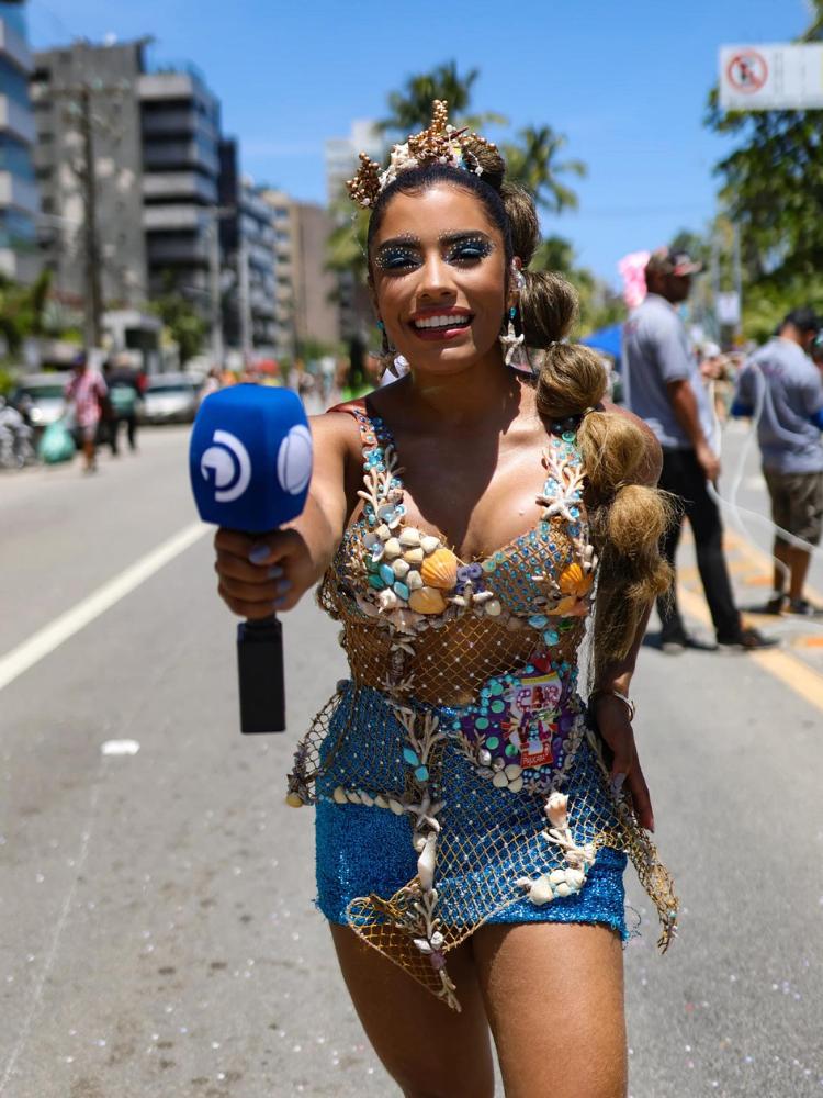 Mulher de pele morena com microfone, top telado com pérolas e conchas, saia azul, conchas no cabelo e pérolas na make