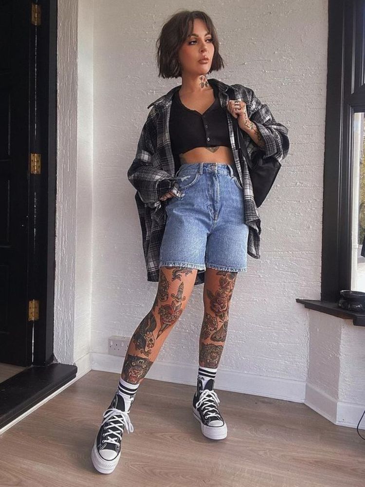 Mulher tatuada usando cropped preto, bermuda jeans, camisa xadrez e all star.