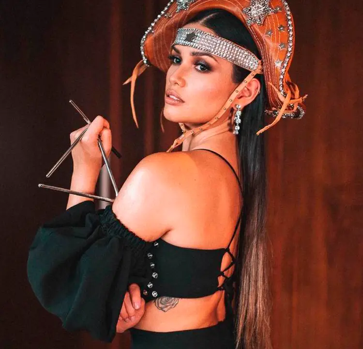 Foto de Juliette Freire de lado, com calça e top pretos e chapéu de cangaceiro, segurando o instrumento triângulo. Dá para ver uma parte da tatuagem dela nas costelas.