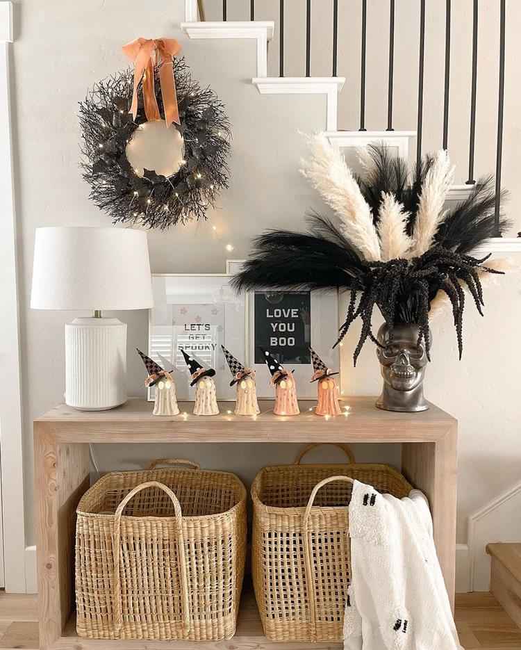 entrada de casa decorada para halloween, mesa de madeira com objetos em formato de fantasma e vaso em formato de caveira, guirlanda seca, quadros, abajur branco e cestos de palha