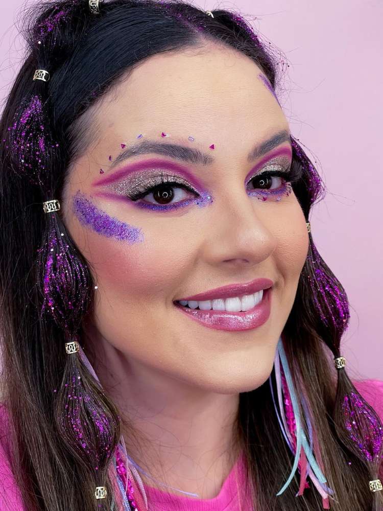 Mulher de pele clara com maquiagem roxa de glitter nos olhos, brilho roxo acima da sobrancelha, delineado roxo e glitter roxo na maça do rosto.