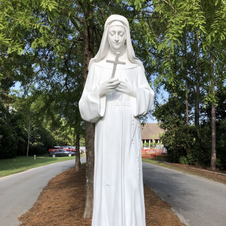 Estátua branca de Santa Rita de Cássia em um jardim arborizado, segurando um pequeno crucifixo contra o peito, simbolizando devoção e serenidade.