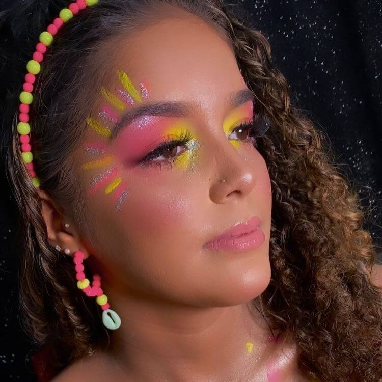 Mulher de pele morena usando maquiagem em rosa e amarelo com raios solares perto da sobrancelha nas mesmas cores e glitter prata 