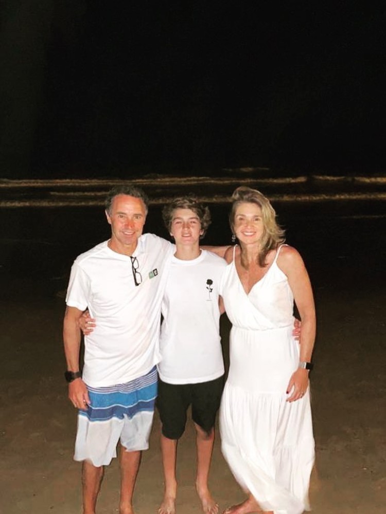 Na praia, João aos 14 anos ao meio usando blusa branca e bermuda preta, abraçando o pai que usa blusa branca e bermuda azul e a mãe que usa vestido branco 
