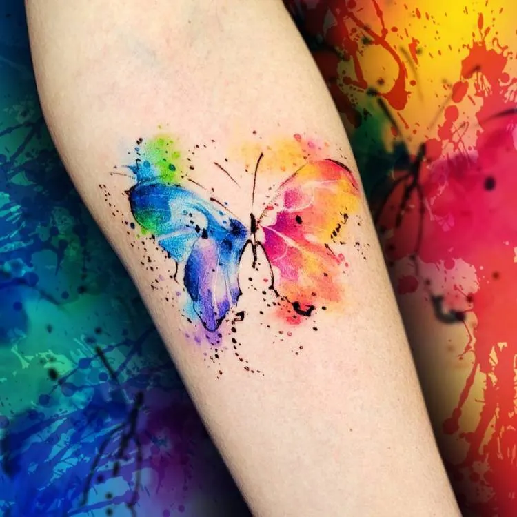 Tatuagem estilo aquarela em tons de azul, verde, rosa e roxo no braço de uma mulher de pele clara. 