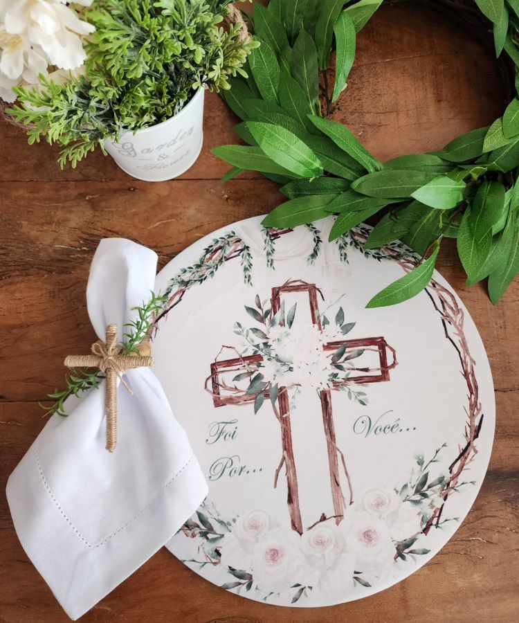 Mesa decorativa com temática religiosa, composta por prato com ilustração de cruz adornada por flores e palavras “Foi Por... Você...”. Guardanapo branco com mini cruz decorativa, guirlanda de folhas verdes e vaso branco com arranjo floral complementam a cena.