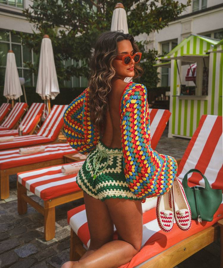 Uma mulher loira de costas, com óculos de sol laranja, usa um conjunto de crochê colorido: um top cropped de manga longa com pontos quadrados coloridos e um shorts listrado verde e branco. Ela está ajoelhada em uma espreguiçadeira de piscina listrada de laranja e branco.