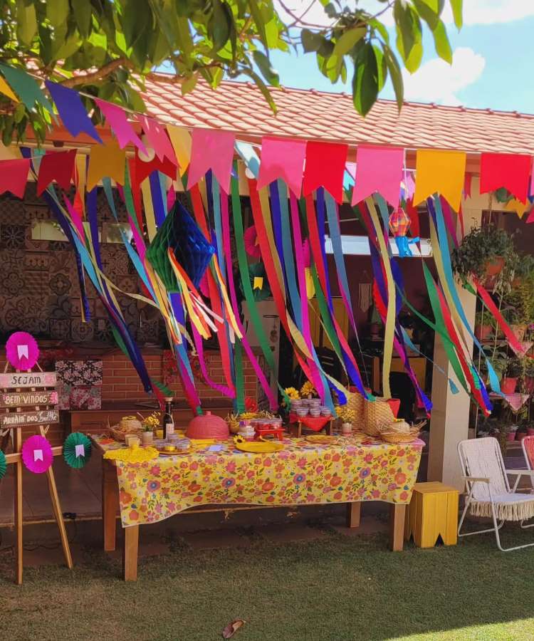 Uma mesa coberta com uma toalha floral amarela, decorada para uma festa junina ao ar livre. Bandeirolas coloridas penduradas e arranjos de fitas complementam o cenário festivo e convidativo.