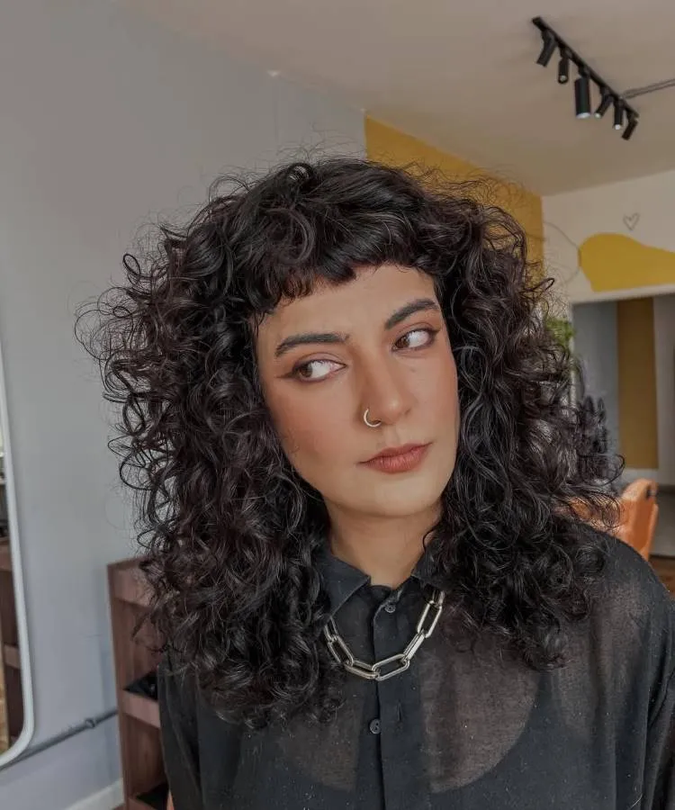 Uma mulher com um corte de cabelo cacheado escuro e franja curta, usando uma camisa preta e um colar de corrente. Ela olha para o lado com um piercing no nariz e maquiagem destacada nos olhos.