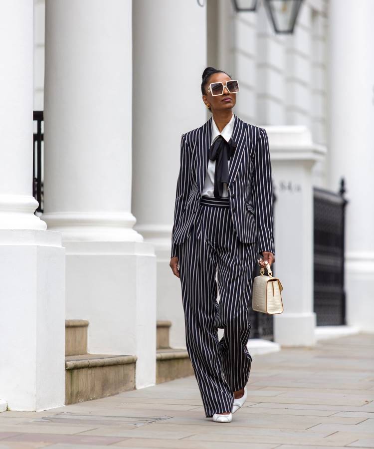 Mulher confiante veste terno risca de giz azul-marinho, camisa branca com laço preto e sapatos brancos. Carrega bolsa estruturada creme e usa óculos de sol grandes. Fundo com colunas brancas clássicas. Elegante inspiração para looks de trabalho sofisticados e modernos.





