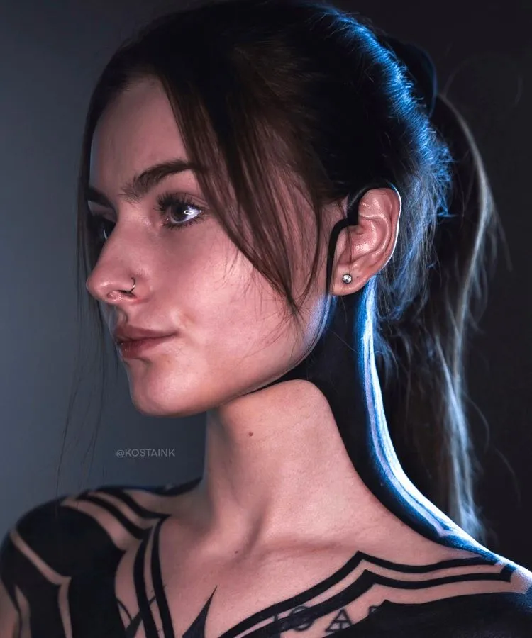 A imagem mostra uma pessoa de perfil com cabelo castanho preso em um rabo de cavalo. Ela tem uma tatuagem no pescoço que é uma faixa preta que se estende até o ombro. A pessoa usa um piercing no nariz e um brinco pequeno na orelha.


