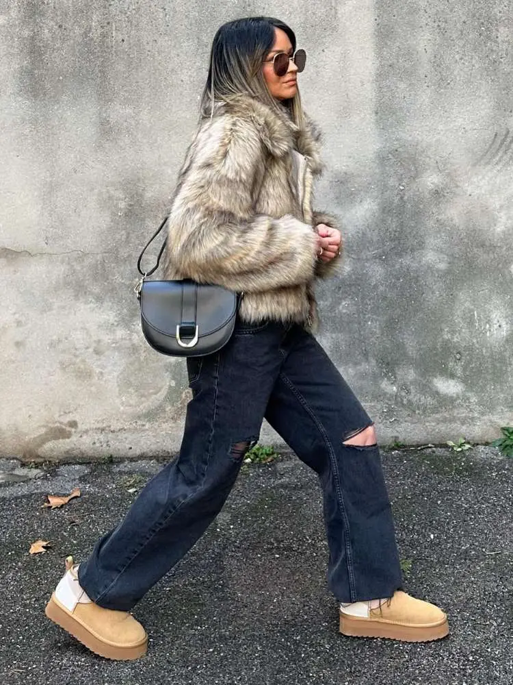 Mulher de pele clara usando casaco de pele bege, calça jeans escura, bolsa de couro preta e ugg boots 