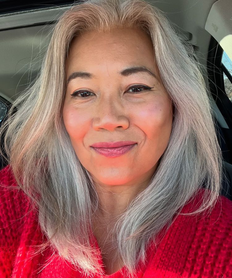 Mulher de cabelos longos e grisalhos com reflexos prateados, usando maquiagem leve e batom rosa. Ela veste um suéter vermelho e está sentada em um carro, em um ambiente iluminado com luz natural suave.