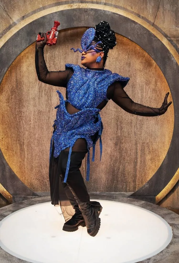Uma pessoa com maquiagem e cabelo elaborados, usando um traje futurista azul brilhante, com botas de cano alto. Ela segura uma arma de brinquedo vermelha e posa em um palco circular, com uma parede cinza atrás.