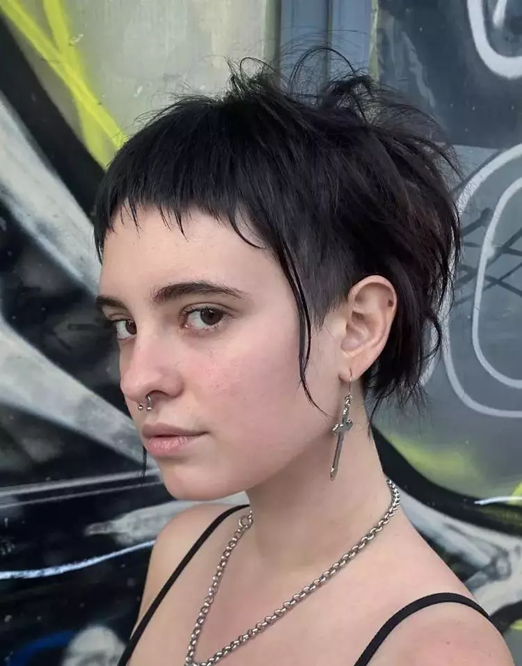 Menina com corte de cabelo curto com microfranja, usando piercing e joias prateadas de cruz.