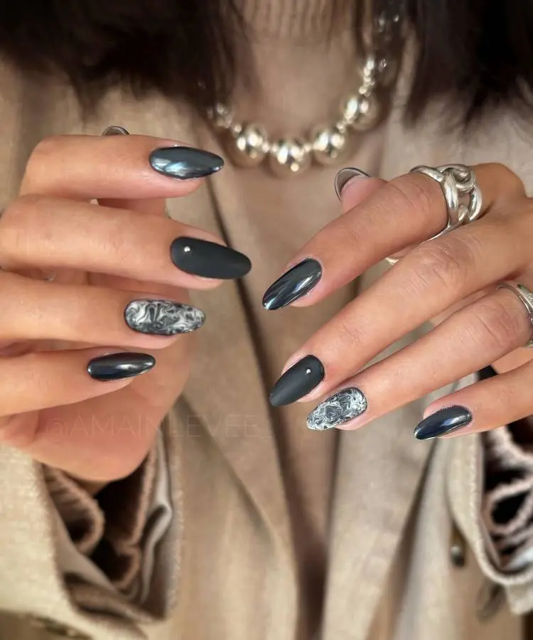 A imagem mostra mãos com unhas em tons de cinza. Algumas unhas têm um acabamento metálico, enquanto outras apresentam um design marmorizado em preto e branco. As unhas são longas e bem cuidadas. A pessoa usa anéis prateados e um colar de pérolas, complementando o visual sofisticado e moderno. O fundo é neutro, destacando as unhas.


