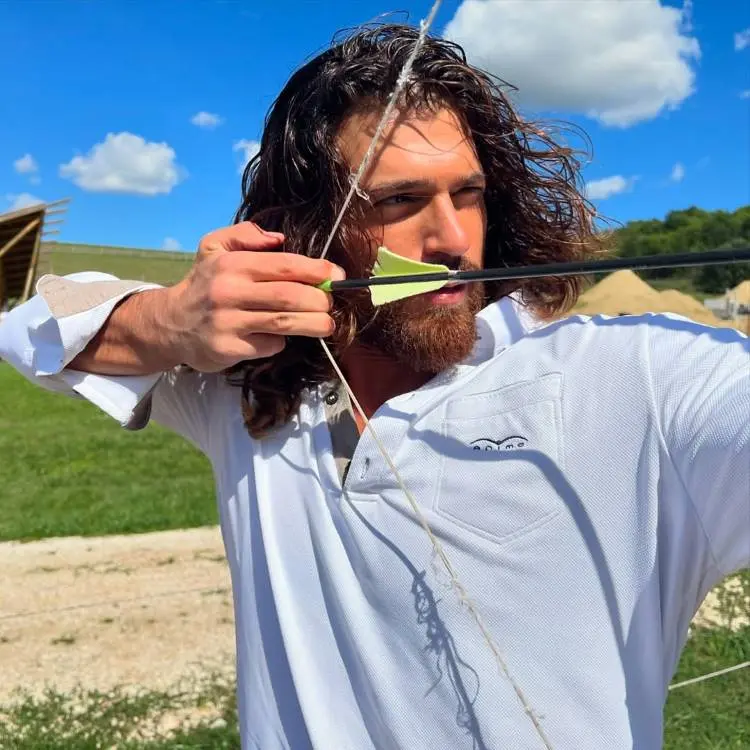 Homem de cabelos longos e barba, vestindo camisa branca, concentra-se enquanto puxa o arco com uma flecha. O cenário ao fundo é ao ar livre, com gramado verde, céu azul e nuvens brancas.