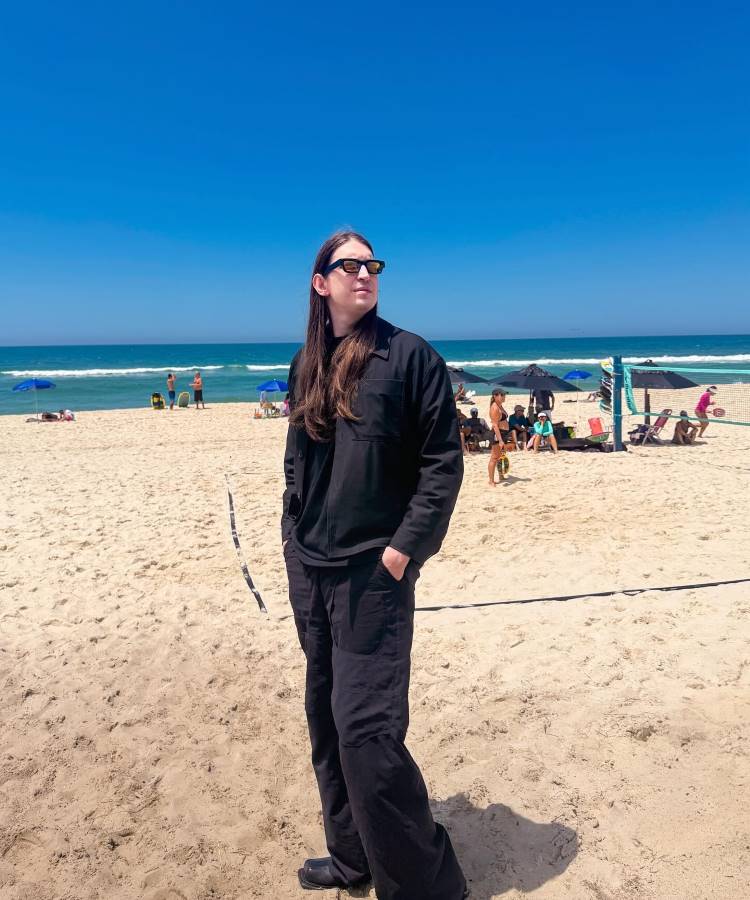 Homem de cabelo comprido e óculos escuros, vestido todo de preto, em pé na areia de uma praia ensolarada. Ele olha para o lado, com a mão no bolso. Pessoas e barracas de praia no fundo.