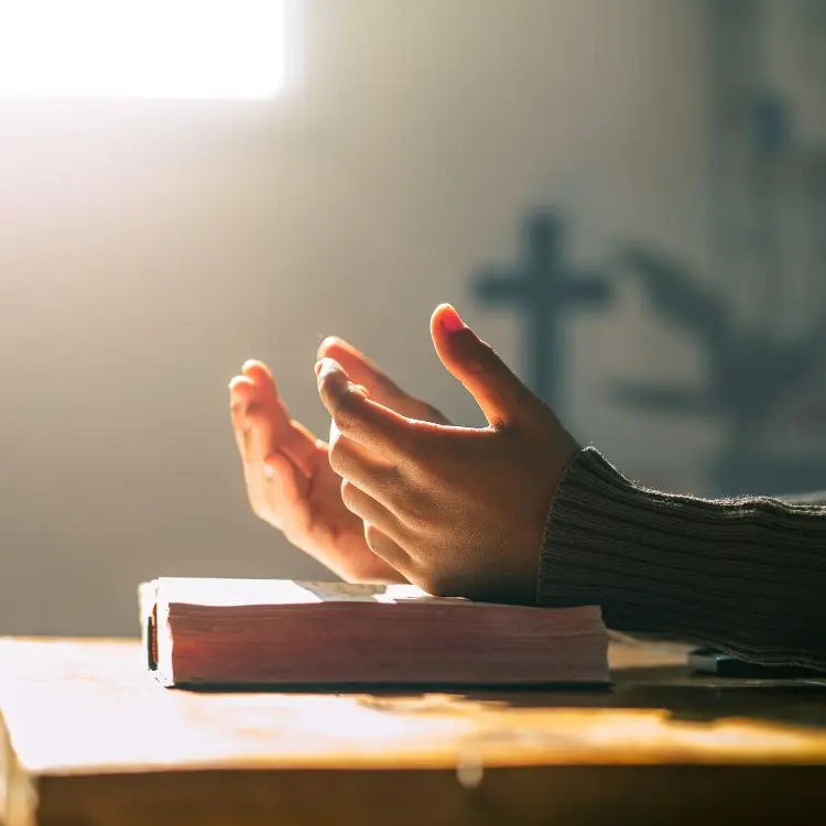 Mãos levantadas em gesto de oração, próximas a uma Bíblia aberta sobre uma mesa de madeira. Ao fundo, luz natural entra pela janela, iluminando suavemente o ambiente que exibe uma cruz desfocada em segundo plano.