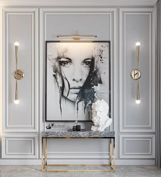 Hall de entrada com quadro em preto e branco, escultura de busto de homem, luminárias, aparador em pedra e dourado e boiserie branco em parede cinza