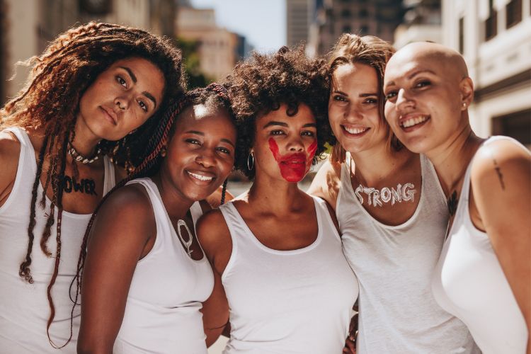Cinco mulheres usando regata branca em protesto feminino. elas estão pintadas com tinta: uma no peito escrito Power, outra com símbolo do feminismo, outra com uma mão na boca e outra escrito strong
