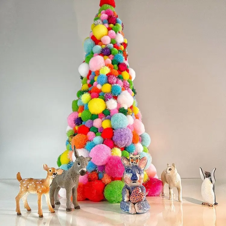Uma árvore de Natal feita de pompons coloridos, cheia de cores vibrantes como rosa, azul, verde e amarelo. Ao redor, cinco pequenas esculturas de animais: dois veados, um coelho, um cavalo e um pinguim, criando uma cena alegre e festiva.