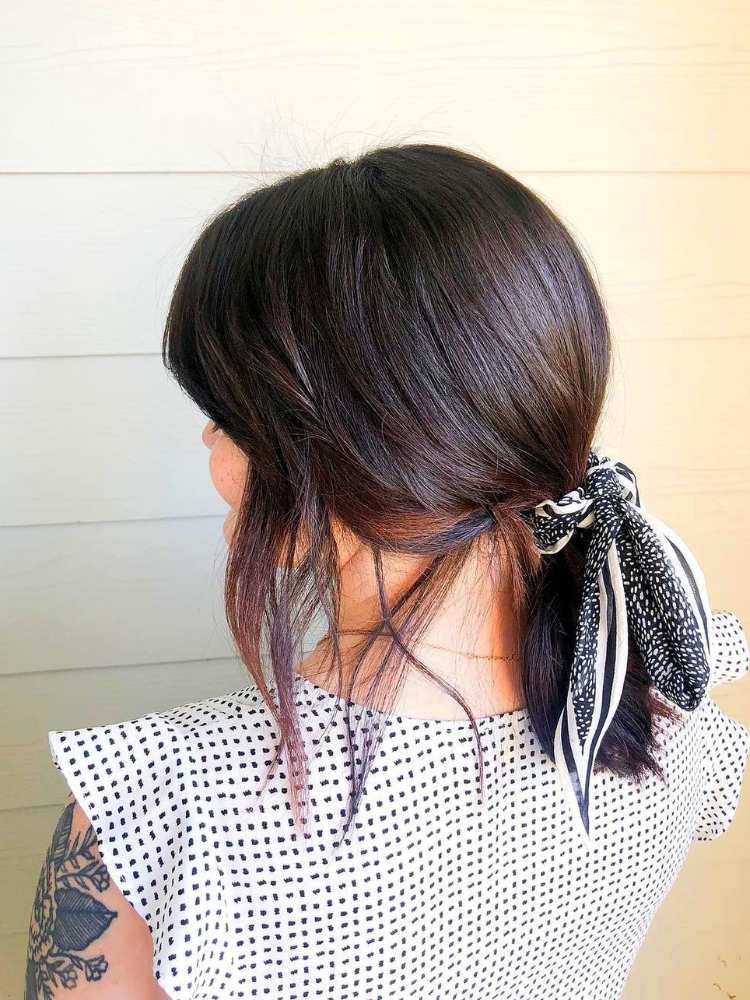 Mulher de pele clara e cabelo escuro de costas, usando blusa preta e branca e rabo de cavalo baixo com lenço branco e preto