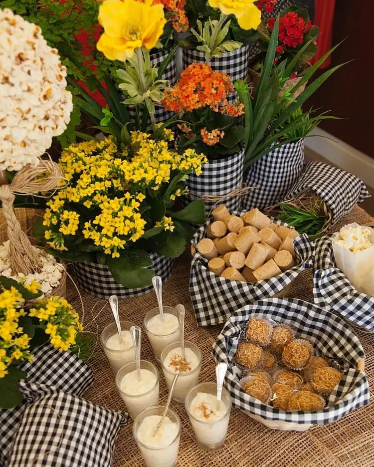 A imagem representa potes, vasos e outras decorações de festa junina enfeitas com tecido xadrez preto e branco. Estão em uma superfície, contém canjica, paçoca, pipoca e cocada