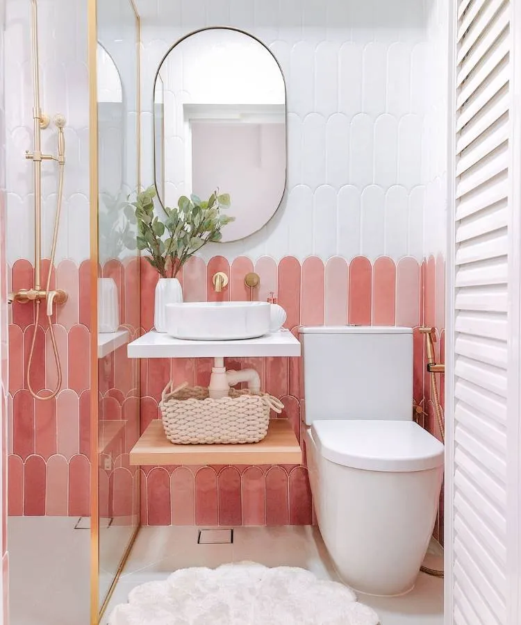 Um banheiro moderno com azulejos de dois tons de rosa pastel, pia de bancada branca com torneira dourada, vaso sanitário branco e chuveiro dourado. Há uma planta verde em um vaso branco e uma cesta de vime sob a pia.

