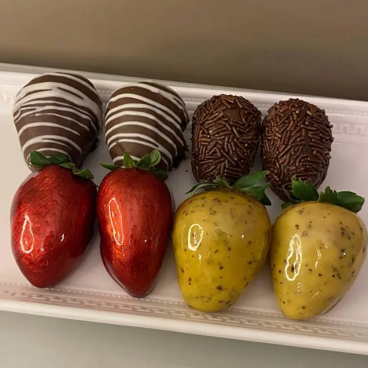 Uma bandeja branca com oito morangos decorados. Quatro são de chocolate, dois com granulado e dois com riscas brancas. Os outros quatro são caramelizados, dois em vermelho metálico e dois em amarelo com pintinhas pretas.