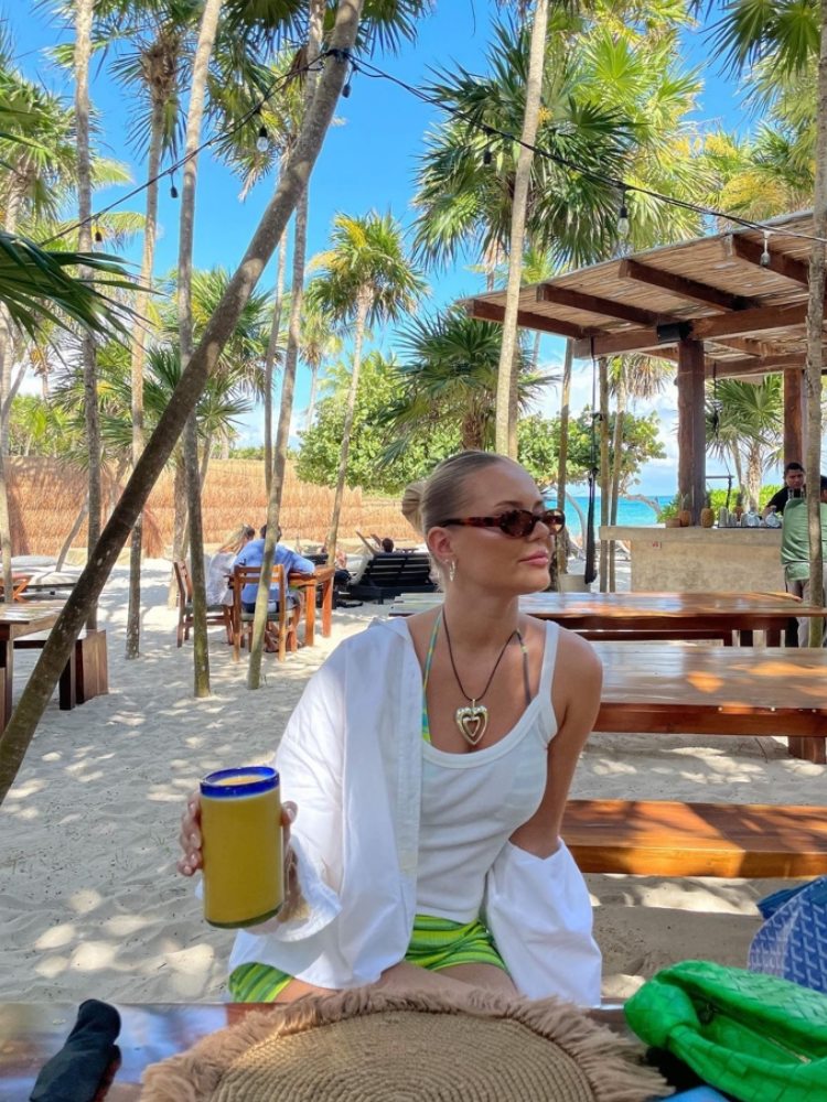 mulher usando regata sob biquini colorido com sobreposição de camisa branca e shorts verde. ela esta de cabelo preso, oculos de sol marrom e colar com pingente de coração.
