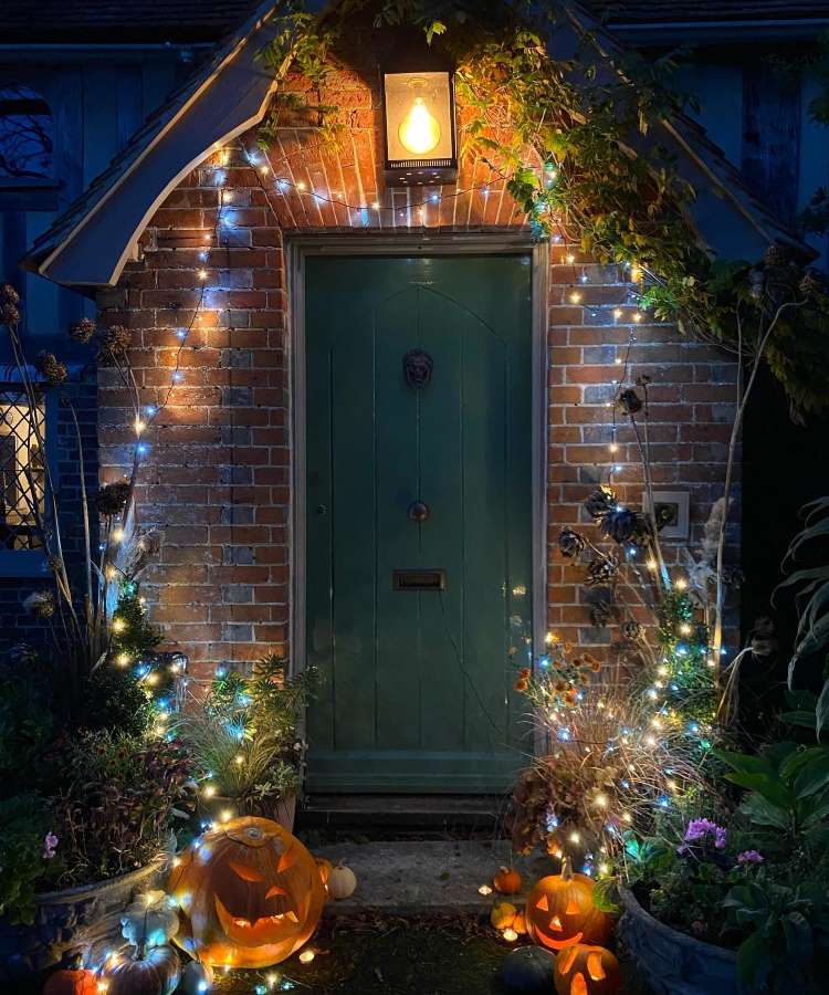 Entrada de casa com tijolos à vista, porta verde, fios de luzes, flores e galhos secos e abóboras iluminadas 