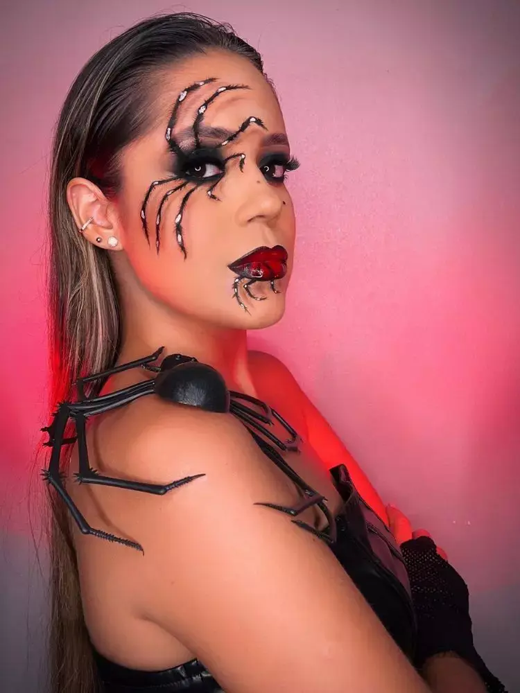 Mulher de pele clara com desenho de aranha no olho e na boca, batom vermelho e aranha de brinquedo no ombro