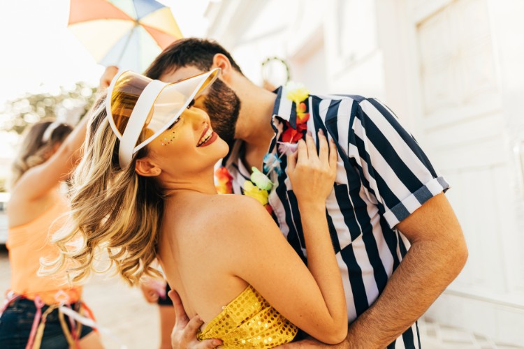 Mulher de pele clara com top dourado e viseira abraçada com homem de pele clara usando camisa listada branca e preta no Carnaval