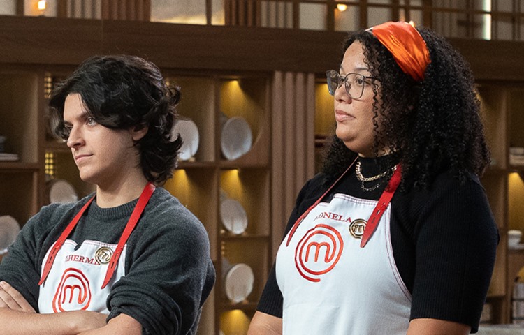 Dois participantes do MasterChef Brasil, um homem de cabelo escuro e uma mulher de óculos e faixa laranja, estão usando aventais brancos. Ambos olham para o lado na cozinha do programa, com um fundo desfocado de prateleiras com pratos.
