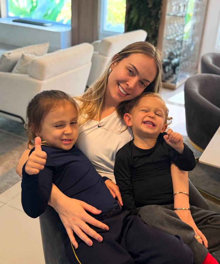 A imagem mostra Tata Estaniecki sentada com seus dois filhos. Tata sorri para a câmera, enquanto a filha faz um sinal de "joinha" e o filho ri com os olhos fechados. O trio está em um ambiente interno, com móveis ao fundo.